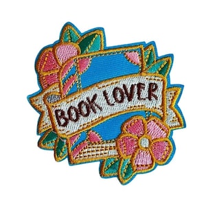 Könnte beinhalten: Gestickter Aufnäher mit blauem Hintergrund und einem Banner mit der Aufschrift "BOOK LOVER" in braunem Text. Der Aufnäher zeigt rosa und gelbe Blumen, grüne Blätter und einen goldenen Rand. Der Aufnäher ist zum Aufbügeln konzipiert.