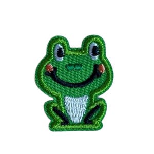 Könnte beinhalten: Gestickter Aufnäher einer lächelnden grünen Frosch mit schwarzen Augen und einem weißen Bauch. Der Frosch hat einen schwarzen Mund und kleine rote Akzente auf den Wangen. Der Aufnäher ist mit dunklerem Grün umrandet.
