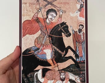 Könnte beinhalten: Eine religiöse Ikone mit einem Mann auf einem schwarzen Pferd mit gekreuzten Schwertern, einer Figur in roten Gewändern und anderen Figuren. Das Kunstwerk hat einen bordeauxfarbenen Rand und wird von einer Hand gehalten.