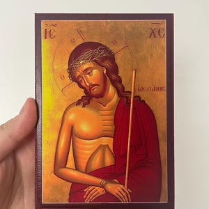 Può includere: Icona religiosa raffigurante una figura con una corona di spine, che indossa una veste rossa e tiene in mano un bastone. L'immagine ha toni oro e marrone, con testo negli angoli superiori. L'icona è su una tela con cornice rosso scuro.