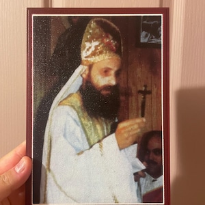 May include: A framed portrait of a religious figure wearing a gold and white headdress and holding a cross. The portrait has a burgundy frame and is set against a neutral background.