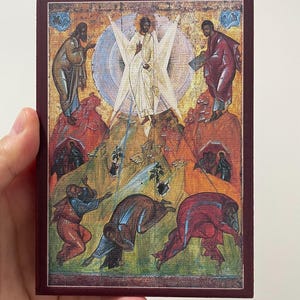 May include: A religious icon featuring a central figure in white robes, surrounded by a bright light. Figures in robes are depicted on the sides and below, set against a colorful, textured background. The icon is framed in a dark red border.