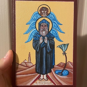 Pode incluir: Ícone religioso com uma figura de longa barba branca e vestes azuis, mãos juntas em oração. Acima, um anjo com asas azuis e uma auréola. O fundo é uma cena desértica com uma torre e uma planta estilizada.