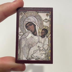 Può includere: Una piccola icona religiosa rettangolare raffigurante la Vergine Maria e il bambino. L'immagine ha uno stile dettagliato e tradizionale con un bordo bordeaux. Le figure sono vestite di bianco e oro, con aureole.