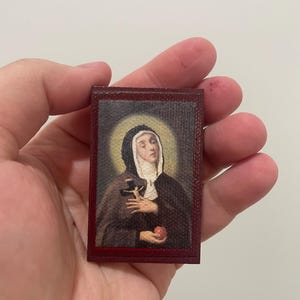 Könnte beinhalten: Ein kleines, rechteckiges religiöses Icon mit dem Porträt einer Nonne. Die Nonne trägt einen schwarzen Habit und eine weiße Haube und hält ein Kreuz und einen kleinen roten Gegenstand. Das Icon hat einen dunkelroten Rahmen.