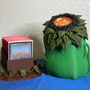 May include: A green, stylized card holder shaped like a plant, with a dark rim and orange center. A brown card holder with red-edged cards and a silver sphere with the text "3D by" are also present. The items are on a blue surface.