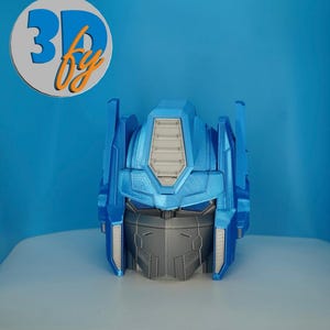 May include: A 3D-printed model of a robot's head, primarily blue with gray and white accents. The head features a detailed design with angular shapes and panels. The logo "3D by" is in a circle in the upper left corner.
