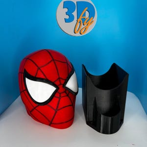 May include: A red and black Spider-Man mask with white eye lenses, next to a black, angular container. The mask has a web pattern. The background is blue with a logo that says "3D fy".