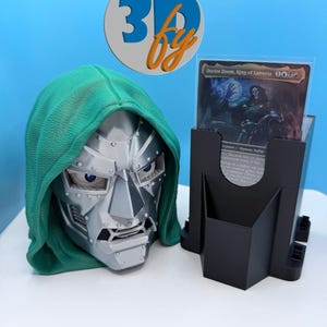 May include: A Doctor Doom bust with a green hood and silver metallic mask, positioned next to a black plastic card display stand holding a Doctor Doom, King of Latveria trading card. The card features text including the name and game stats.