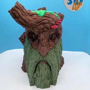 May include: A 3D-printed sculpture of a tree-like creature with a detailed face and beard. The sculpture features a brown head with green and red accents, and a long green beard. The logo "3D by" is in the upper right corner.