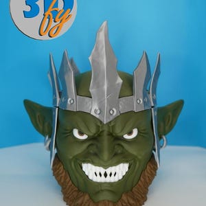 May include: A green goblin-like mask with a silver crown and a brown beard. The mask has a menacing expression with sharp white teeth. The image includes the text "3D by" in a circular logo.