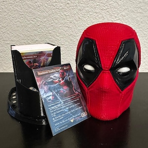 May include: A black and red 3D printed Deadpool head figurine with a black and white mask. The figurine is sitting on a black surface next to a black card holder with a trading card featuring Deadpool. The card reads "Deadpool, Trading Card" and has a red and black illustration of Deadpool.