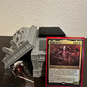 May include: A red-bordered Magic: The Gathering card featuring Edgar Markov, a Vampire Knight, next to a gray 3D-printed card holder. The card depicts a group of vampires. A small figurine of a vampire with a sword is in front of the holder.