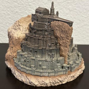 May include: A 3D printed model of a stone castle on a rocky base. The castle is detailed with towers, walls, and windows. The model is painted in shades of gray and brown.