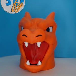 May include: An orange 3D-printed figure of a dragon's head with an open mouth, revealing a red interior and white teeth. The figure has blue eyes and a logo in the upper left corner. The background is a gradient of blue and white.