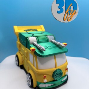 May include: A 3D-printed yellow and green toy van, reminiscent of the Teenage Mutant Ninja Turtles' vehicle. The van features a green top with silver details, a green front bumper, and the word "TURTLES" on the front. The wheels are silver.
