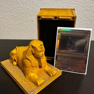 May include: A gold-colored sphinx figurine with a card box behind it. The card box is open and has a Magic: The Gathering card inside. The card is titled "Khephra, the Scarab God".