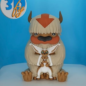 May include: A detailed figurine of Appa from Avatar: The Last Airbender, featuring a tan body, orange arrow, and a smaller figure. The figurine has a light blue background and the text "3D by" in a circle.