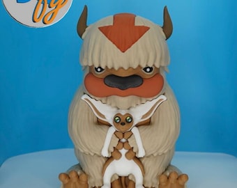May include: A detailed figurine of Appa from Avatar: The Last Airbender, featuring a tan body, orange arrow, and a smaller figure. The figurine has a light blue background and the text "3D by" in a circle.