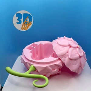 May include: A pink, flower-shaped container with an open lid, revealing an interior with compartments. A green, curved handle extends from the base. The container is set against a blue background, with the text "3D by" in a circle.