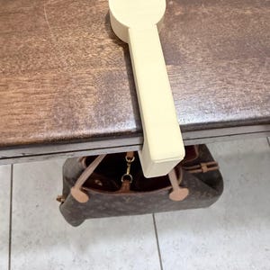 May include: A light beige wooden spoon rest with a circular top and rectangular handle. The spoon rest is resting on a dark brown wooden surface. A brown handbag is visible below the surface.