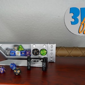 May include: A silver-colored, sword-shaped dice holder with a brown handle. The holder contains various colorful dice. The handle has a stitched pattern. The logo "3D by" is in the upper right corner.