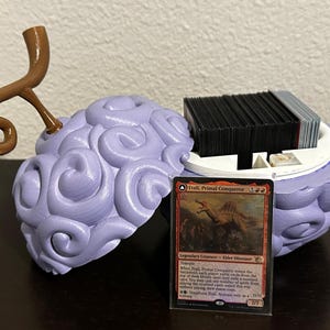 May include: A purple and brown 3D printed card holder shaped like a fruit. The holder is open and a Magic: The Gathering card is visible. The card is titled "Etali, Primal Conqueror" and features a dinosaur.