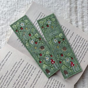 May include: Two green felt bookmarks with embroidered designs of strawberries, flowers, mushrooms, and the name "Ellie". The bookmarks are rectangular and rest on the open pages of a book.