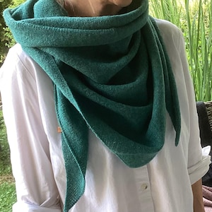 Könnte beinhalten: Ein teal-grüner, dreieckiger Schal, der über einem weißen Hemd drapiert ist. Der Schal besteht aus einem strukturierten Stoff und ist um Hals und Schultern gewickelt. Die Enden des Schals hängen herunter und erzeugen einen geschichteten Effekt. Der Hintergrund zeigt grünes Laub.