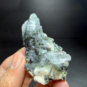 May include: A raw, natural mineral specimen with a mix of colors. The piece features a clear, crystalline structure with shades of green and white, and a dark, textured matrix. The mineral is held in a hand.