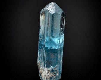 Puede incluir: Un cristal de aguamarina azul claro translúcido con forma prismática hexagonal. El mineral presenta una terminación afilada y facetada en la parte superior y una base rugosa y texturizada con inclusiones minerales blancas en los bordes.