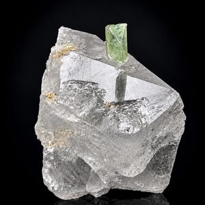 Puede incluir: Un grupo de cristales de cuarzo transparentes y multifacéticos con un pequeño cristal prismático de color verde claro que sobresale de la parte superior. El cuarzo muestra una mezcla de facetas lisas y transparentes y áreas opacas texturizadas con pequeñas inclusiones doradas.