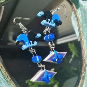 Könnte beinhalten: Ohrringe zum Aufhängen mit einem skurrilen Design. Jeder Ohrring hat einen schwarzen und blauen hutartigen Anhänger, blaue Perlen und eine geometrische Form mit blauen Akzenten. Die Ohrringe sind an silberfarbenen Haken aufgehängt.