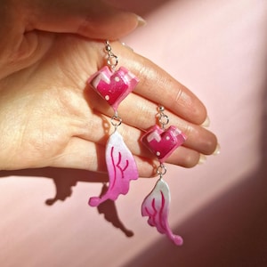 Puede incluir: Pendientes colgantes con una parte superior en forma de corazón y una parte inferior en forma de ala. Los pendientes son de color rosa y blanco, con herrajes de metal plateado. Los pendientes están sostenidos por una mano sobre un fondo rosa.