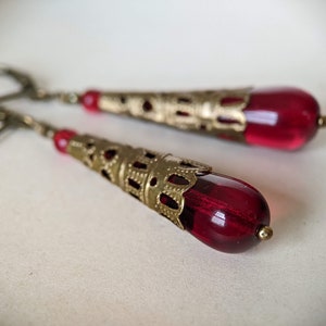 May include: A pair of dangle earrings featuring teardrop-shaped, deep red glass beads. The beads are encased in ornate, gold-colored metal filigree cones. The earrings have a vintage or antique aesthetic, with a lever-back closure.