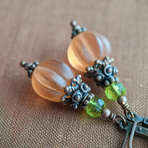 May include: A pair of handcrafted earrings featuring peach-colored, ribbed glass beads and small, bright green beads. The earrings have ornate, antique-style metalwork and a bronze-colored hook closure. The jewelry is set against a neutral brown backdrop.