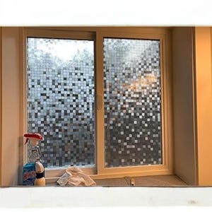 May include: A window with a decorative film applied, featuring a mosaic-like pattern of small, square tiles in shades of gray and white. The window is set in a light-colored wooden frame. Cleaning supplies are visible on the windowsill.
