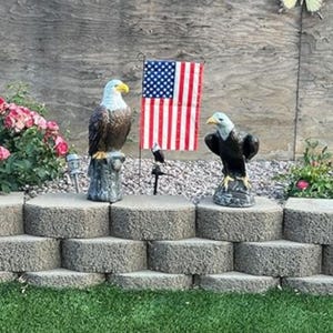 May include: Two eagle statues with a miniature American flag in a garden setting. One eagle is perched on a tree stump, the other on a rock base. The flag has red and white stripes and a blue field with white stars.