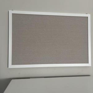 Puede incluir: Un tabl&oacute;n de anuncios rectangular con marco blanco. La superficie del tablero es de color gris neutro texturizado. Est&aacute; montado en una pared gris claro. El tablero est&aacute; vac&iacute;o, listo para notas o fotos.