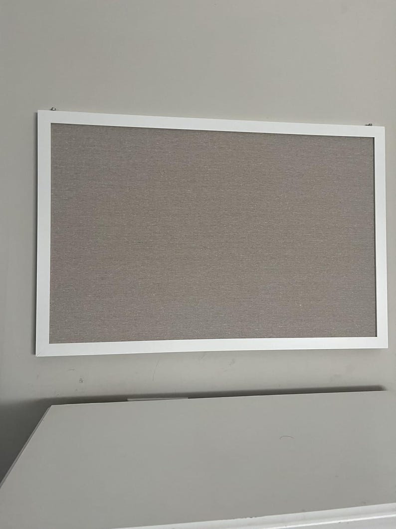 Puede incluir: Un tabl&oacute;n de anuncios rectangular con marco blanco. La superficie del tablero es de color gris neutro texturizado. Est&aacute; montado en una pared gris claro. El tablero est&aacute; vac&iacute;o, listo para notas o fotos.