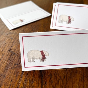 May include: Three white place cards with red borders and a small illustration of a polar bear wearing a red and black plaid scarf.