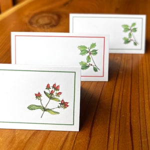 May include: Three white place cards with green borders and different floral illustrations. The top card has a green holly sprig with red berries. The middle card has a red holly sprig with green berries. The bottom card has a green sprig with red berries.
