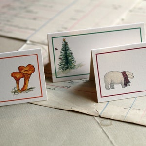 May include: Three white place cards with red, green, and orange borders. Each card features a different illustration: a mushroom, a Christmas tree, and a polar bear wearing a red and black scarf.