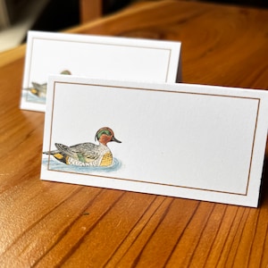 May include: Two white place cards with a gold border. The top card has a simple design. The bottom card features a hand-drawn illustration of a green-headed duck swimming in water.