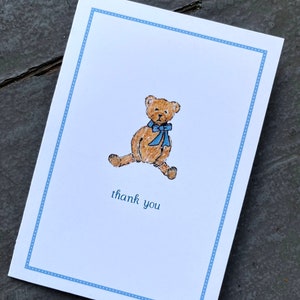 May include: A white card with a blue dotted border. A brown teddy bear with a blue bow is in the center of the card. The text "thank you" is printed below the bear.