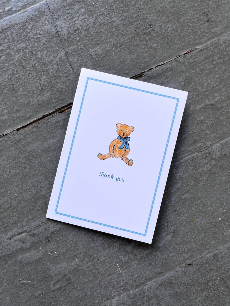 May include: A white thank you card with a blue border. A brown teddy bear with a blue bow is sitting on the card. The text "thank you" is printed on the card.