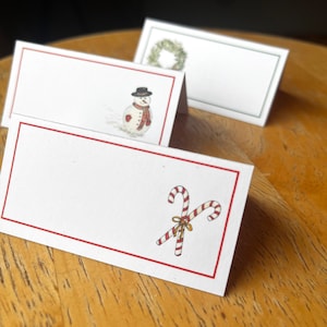 May include: Three white place cards with red borders. The top card has a green wreath, the middle card has a snowman, and the bottom card has two candy canes.