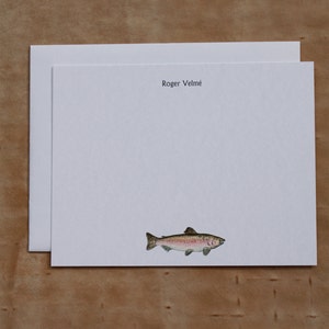 May include: White stationery with the name "Roger Velmé" printed at the top. A watercolor illustration of a fish is at the bottom. The stationery is on a light brown wooden surface.