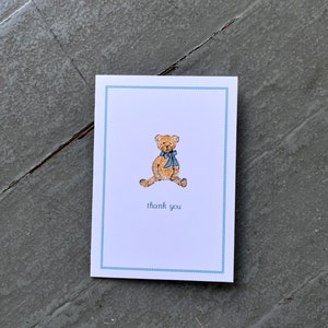 May include: A white card with a blue border and a small illustration of a brown teddy bear wearing a blue bow. The text "thank you" is printed below the bear.