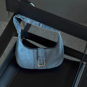 Op de afbeelding: Een lichtblauwe denim schoudertas met een gebogen vorm. De tas heeft een gespsluiting met een goudkleurige metalen gesp. De tas ligt op een zwarte stoel.